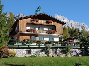 A two-story wooden building with balconies, a stone facade, and a mountain backdrop. - Hotel Villa Gaia (Cortina d'Ampezzo)