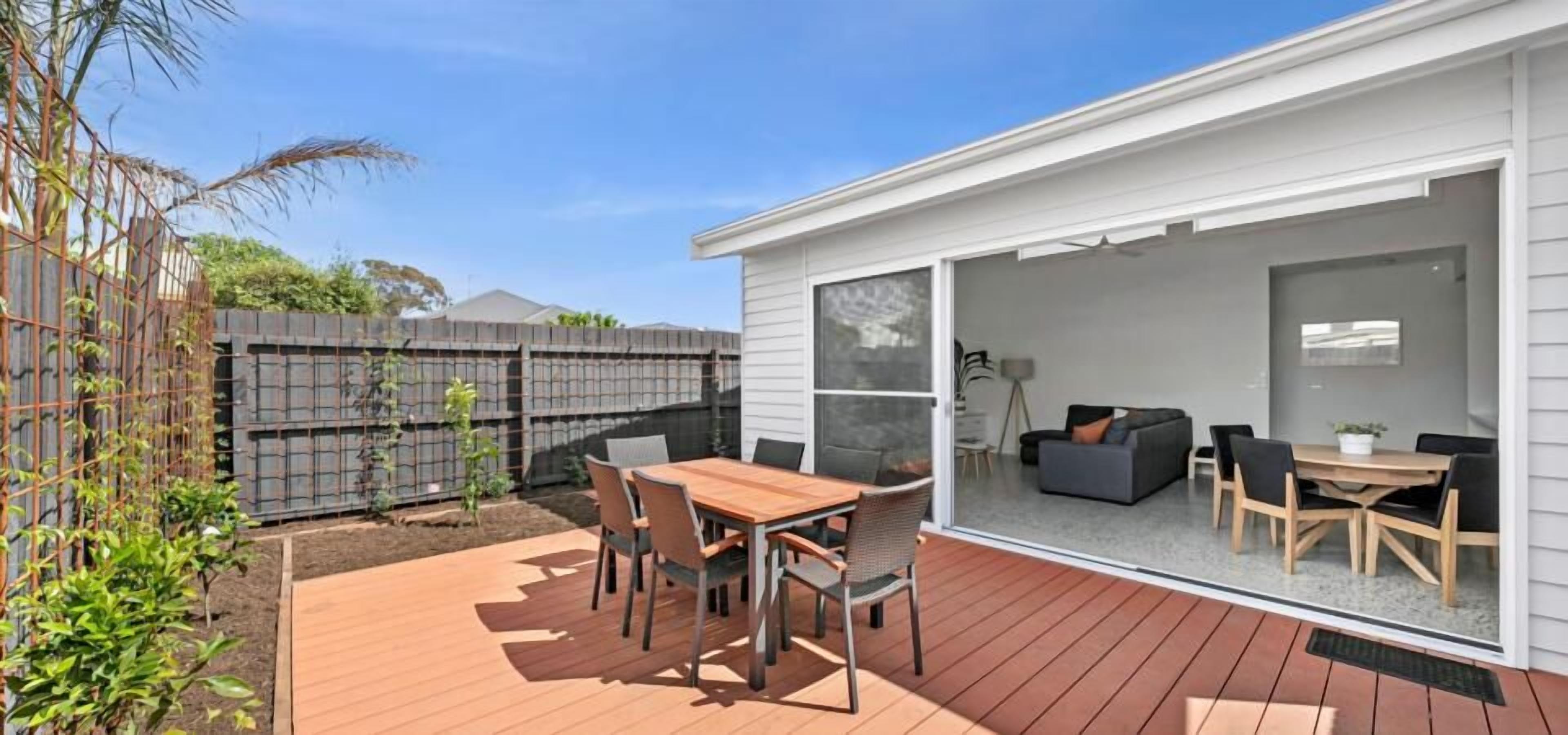 Eine moderne Terrasse mit Holzdeck, einem Esstisch mit Stühlen und einer Glastür, die zu einem Innenbereich mit einer Couch und einem Couchtisch führt.