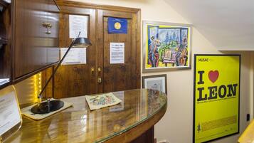 Un área de recepción con un escritorio de madera, una lámpara y varios documentos. Hay obras de arte enmarcadas en la pared y un cartel con texto.