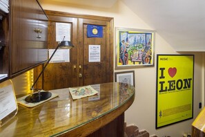 Un área de recepción con un escritorio de madera, una lámpara y varios documentos. Hay obras de arte enmarcadas en la pared y un cartel con texto.