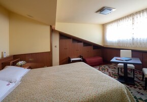 Ein Hotelzimmer mit einem Bett, einem Schreibtisch mit Stuhl, einem kleinen Tisch, einer roten Bank und einem Kleiderschrank.