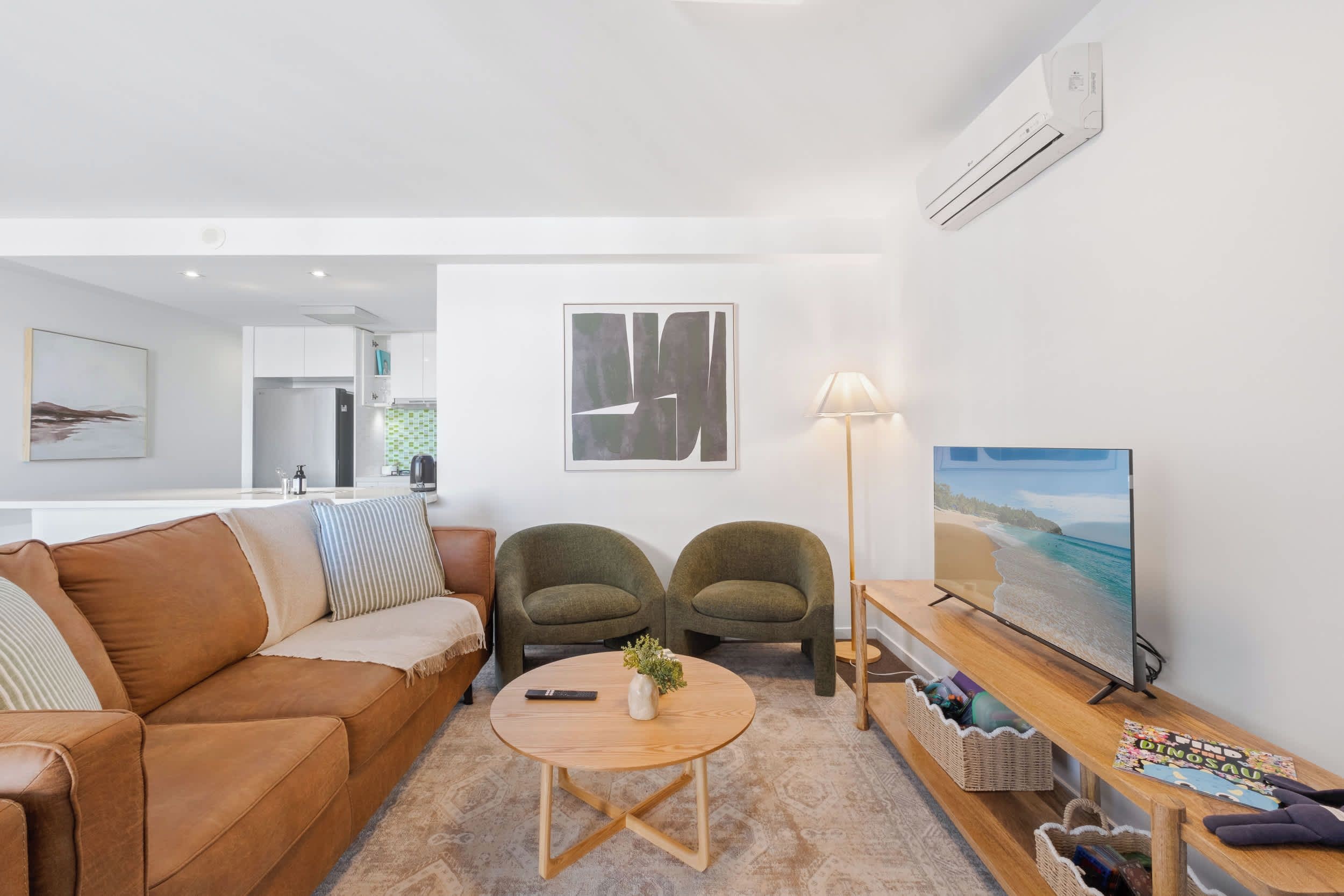 A modern living room with a brown leather sofa, two armchairs, a coffee table with a vase, a floor lamp, and a TV on a wooden stand.