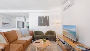 A modern living room with a brown leather sofa, two armchairs, a coffee table with a vase, a floor lamp, and a TV on a wooden stand.