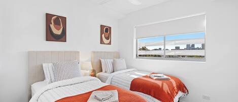 A modern hotel room with two beds, a city view, ceiling fan, and artwork on the wall.