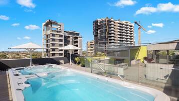 A rooftop pool area with a hot tub, umbrellas, and a view of surrounding buildings.