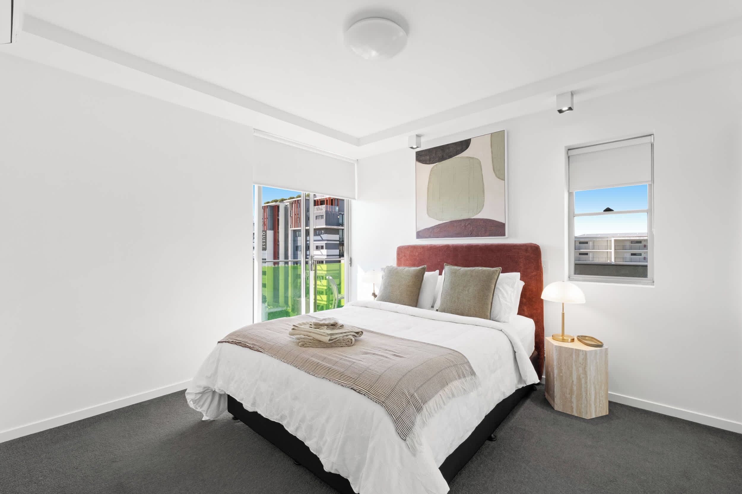 A modern bedroom with a large bed, a nightstand, a lamp, and a view of a building outside.