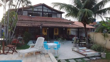 Uma casa de dois andares com telhado de telhas, um pátio com mobiliário de exterior e um jardim com palmeiras.
