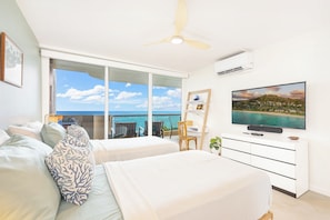Quarto com duas camas, uma escrivaninha, uma cadeira, televisão e vista para o oceano.