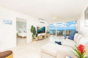 Uma sala de estar moderna com uma televisão grande, um sofá, área de jantar com cadeiras e uma varanda com vista para o mar.