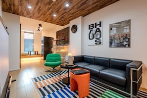 Ein modernes Wohnzimmer mit einem schwarzen Ledersofa, einem grünen Sessel und einem kleinen orangefarbenen Hocker. Die Decke besteht aus Holz und auf dem Boden liegt ein Streifen Teppich.