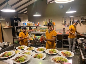 Cozinha com chefs preparando alimentos, bancadas de aço inoxidável e diversos utensílios de cozinha.