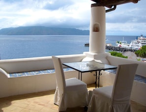 A balcony with a glass table and two chairs, overlooking a body of water and a distant island. - Hotel Santa Marina Antica Foresteria (Santa Marina Salina)