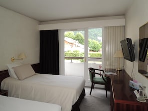 Quarto de hotel com duas camas, uma escrivaninha, uma cadeira, televisão e varanda com vista para o verde.