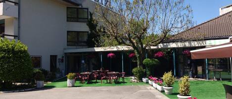 A hotel with a covered outdoor seating area, a garden with potted plants, and a clear sky.