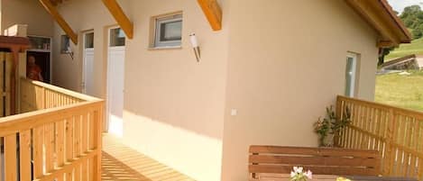 Una terraza de madera con una mesa, bancos, un ventanal y una puerta que da a un espacio interior.
