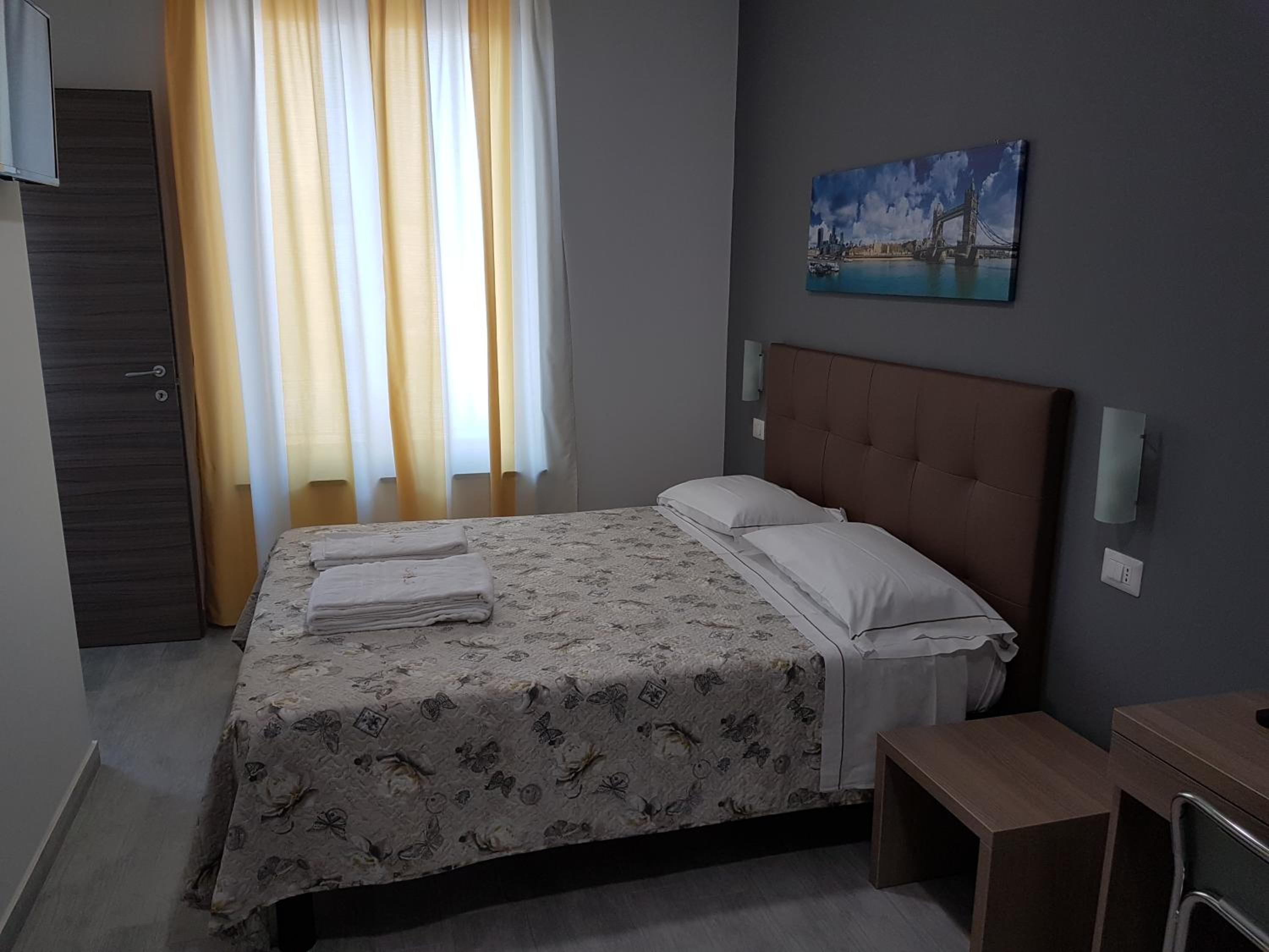 A hotel room with a bed, a desk, a chair, a painting on the wall, and a window with curtains.