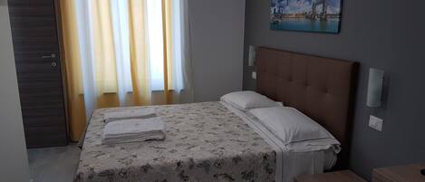 A hotel room with a bed, a desk, a chair, a painting on the wall, and a window with curtains.