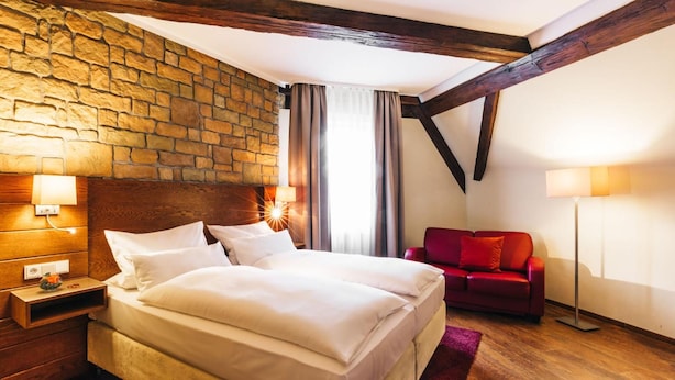 A hotel room with a stone wall, wooden beams, a bed with white linens, a red sofa, and a floor lamp. - Amtshaus Freinsheim (Freinsheim)