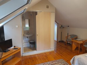 A compact room with a sloped ceiling, a wooden desk, a chair, a small table, and a bed.