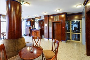 A hotel reception area with a reception desk, chairs, a round table with a glass, and a large pillar. - Nadejda Hotel (Sofia)