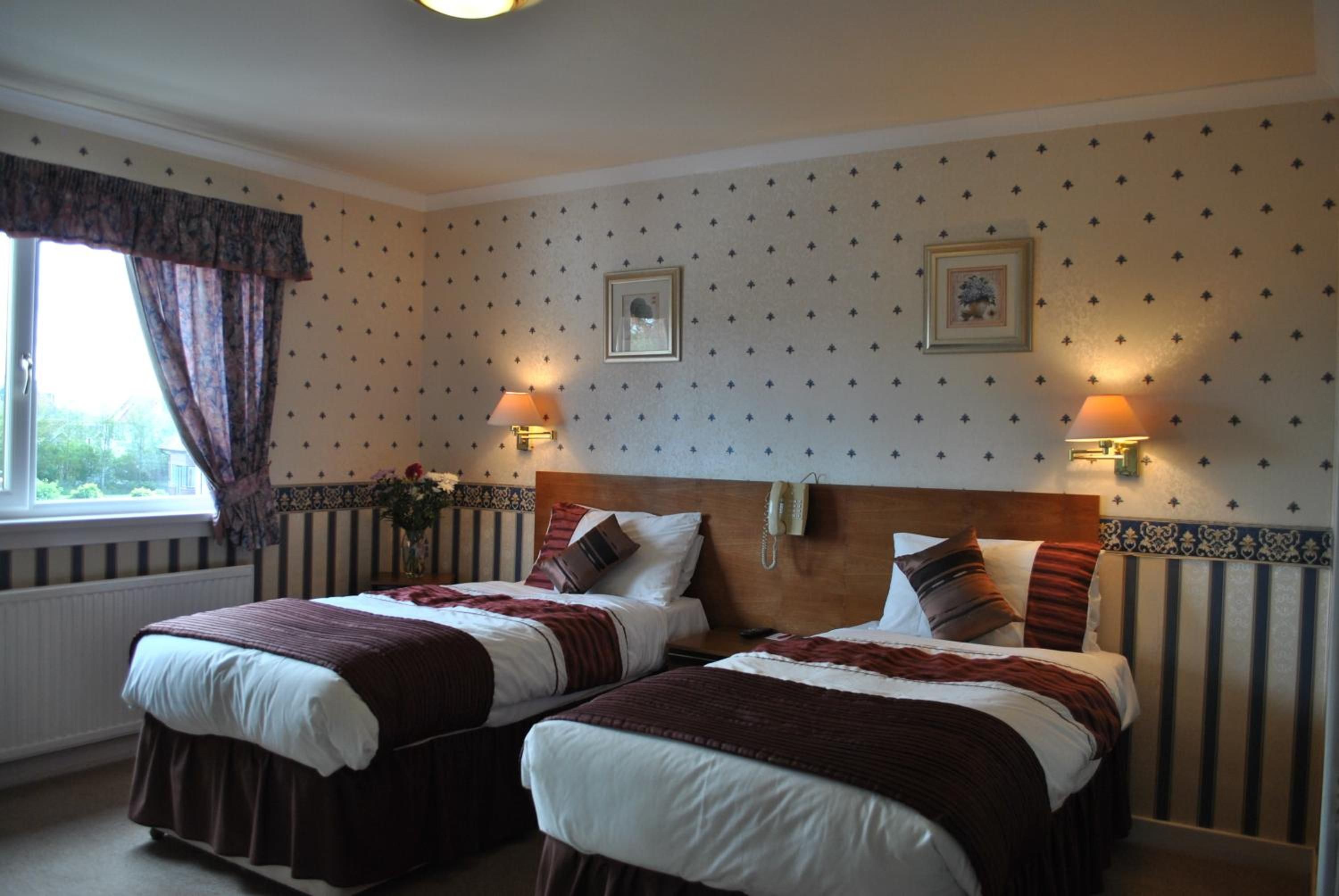 A hotel room with two beds, a window with curtains, wall-mounted lamps, and framed pictures.