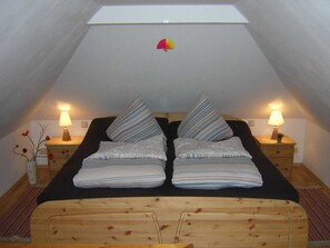 Ein Zimmer mit schrägem Dach, zwei Nachttische mit Lampen, ein Bett mit gefalteter Bettwäsche und ein kleines dekoratives Element an der Decke.
