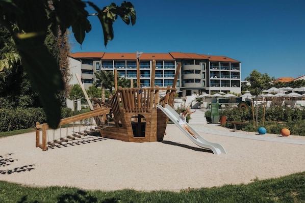 Un parc infantil amb tobogan i estructura per escalar, envoltat d'una zona de sorra i un edifici de diversos pisos al fons.