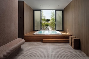 Un baño moderno con una bañera de madera, un banco y vistas a un jardín a través de amplios ventanales.