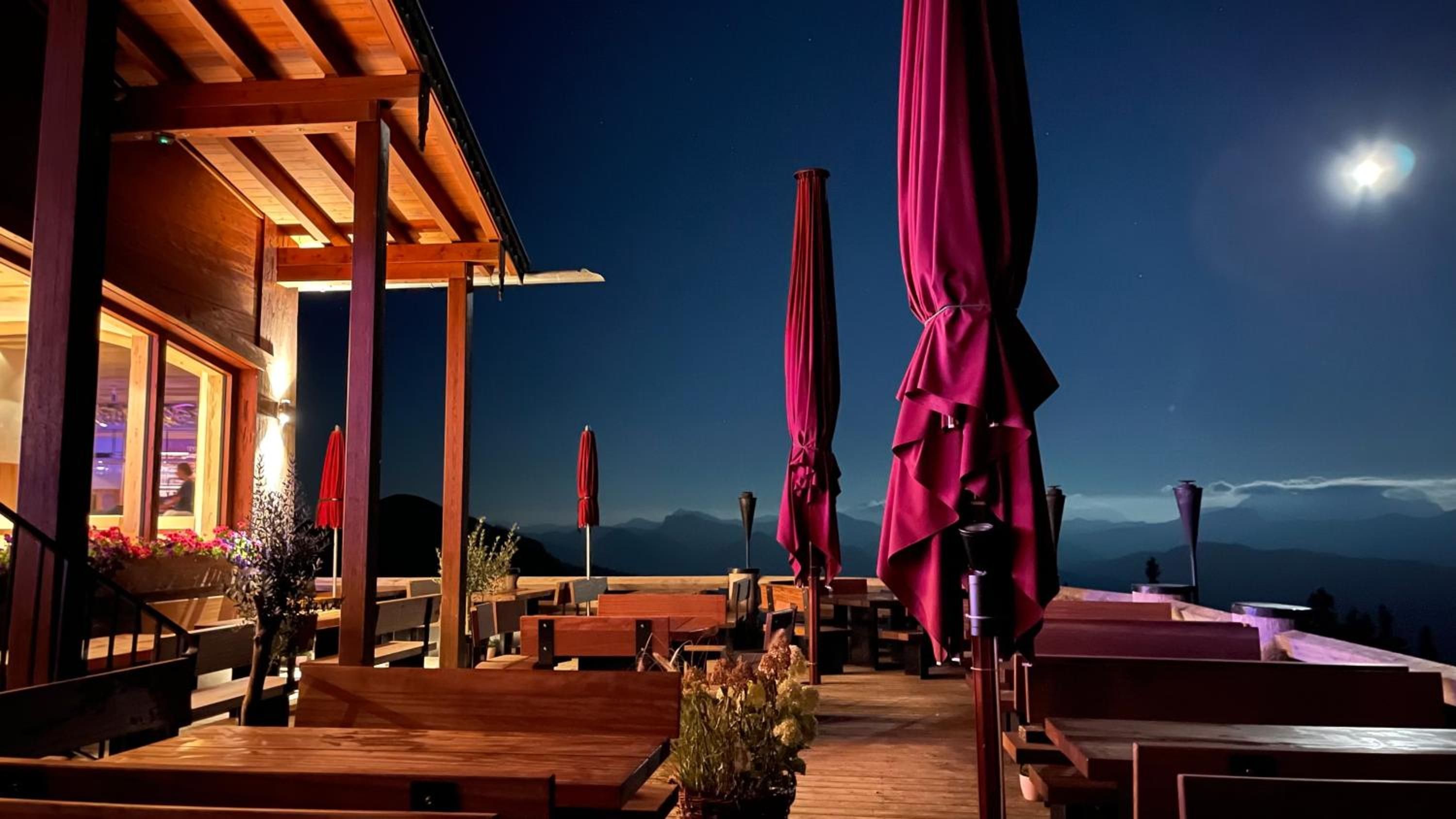 Un espace extérieur aménagé avec des tables et des chaises en bois, des parasols rouges et une vue sur les montagnes sous un ciel nocturne.