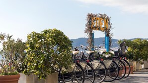 Eine Reihe von Fahrrädern, die in einem dafür vorgesehenen Fahrradständer geparkt sind, mit Blumentöpfen und Blick auf das Meer im Hintergrund.