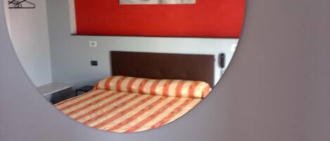 Ein Zimmer mit einem Bett, einer roten Wand und einem Spiegel, der Bett und Wand reflektiert.