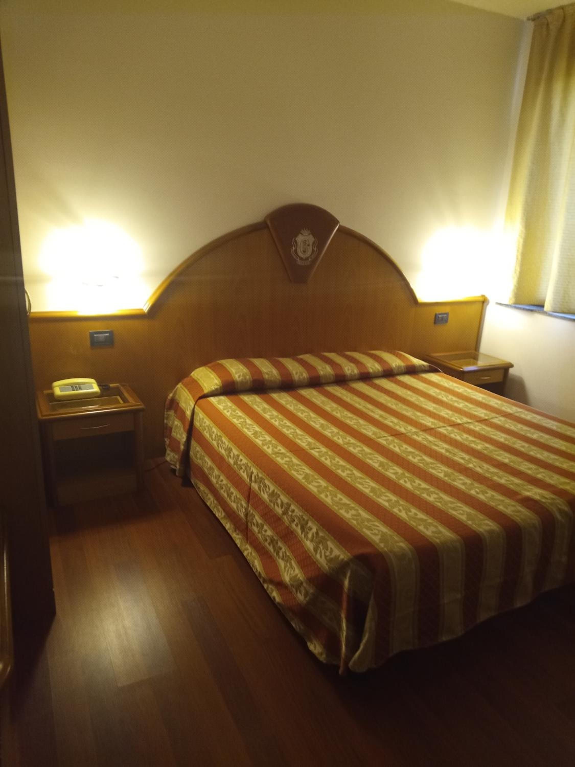 Habitació d'hotel amb un capçal de llit de fusta, un llit amb roba de llit a ratlles, una taula de nit amb una làmpada i una estació de trucades amb un telèfon.
