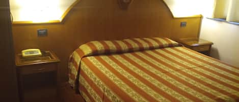 Habitació d'hotel amb un capçal de llit de fusta, un llit amb roba de llit a ratlles, una taula de nit amb una làmpada i una estació de trucades amb un telèfon.