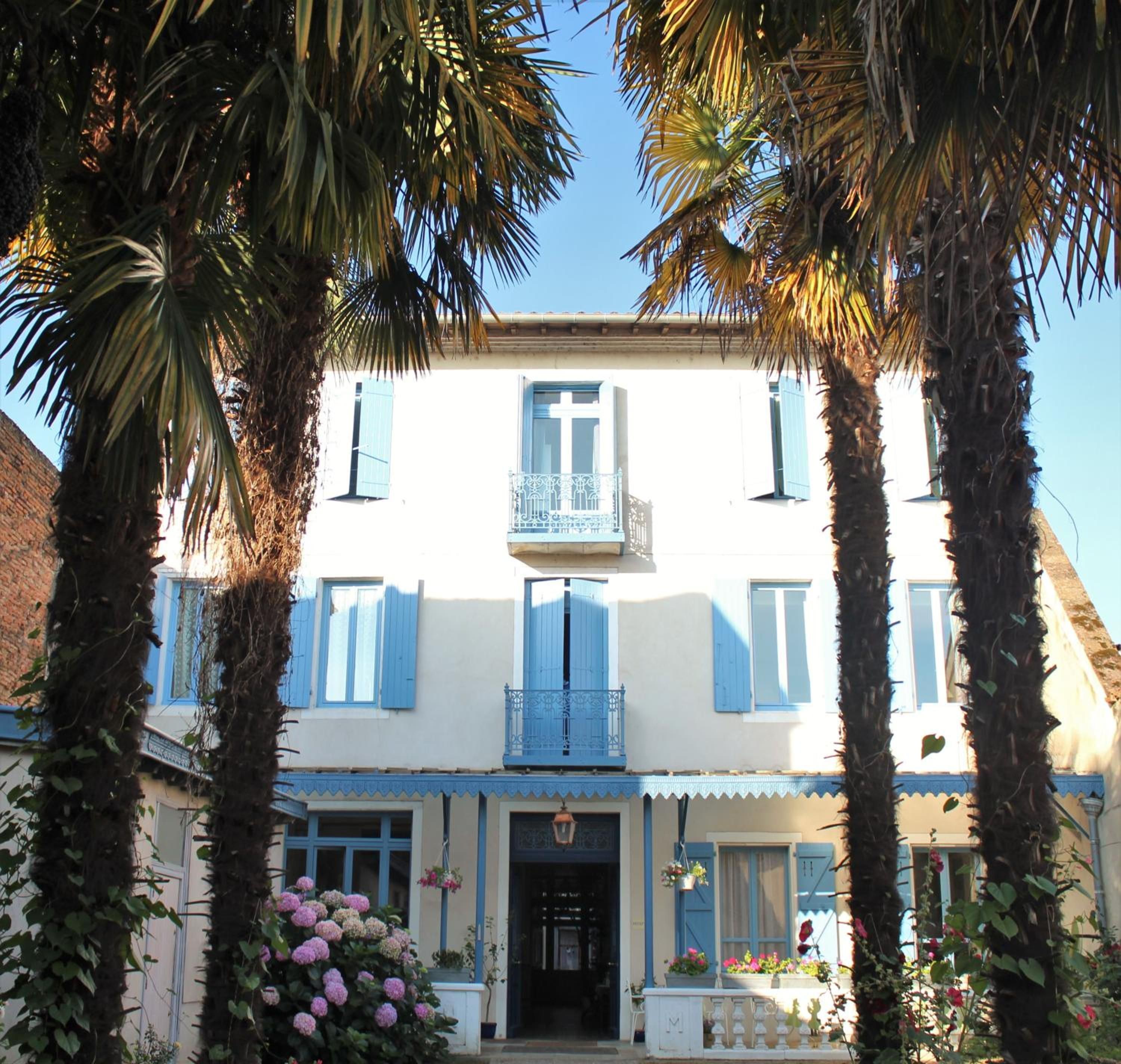 A two-story building with blue shutters, a balcony with a wrought iron railing, and a flower box with pink flowers.