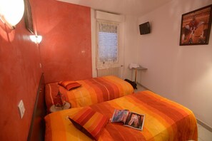 Een kamer met twee bedden, een radiator, een raam met zonwering, een wandlamp, een fotolijst en een tijdschrift op het bed.