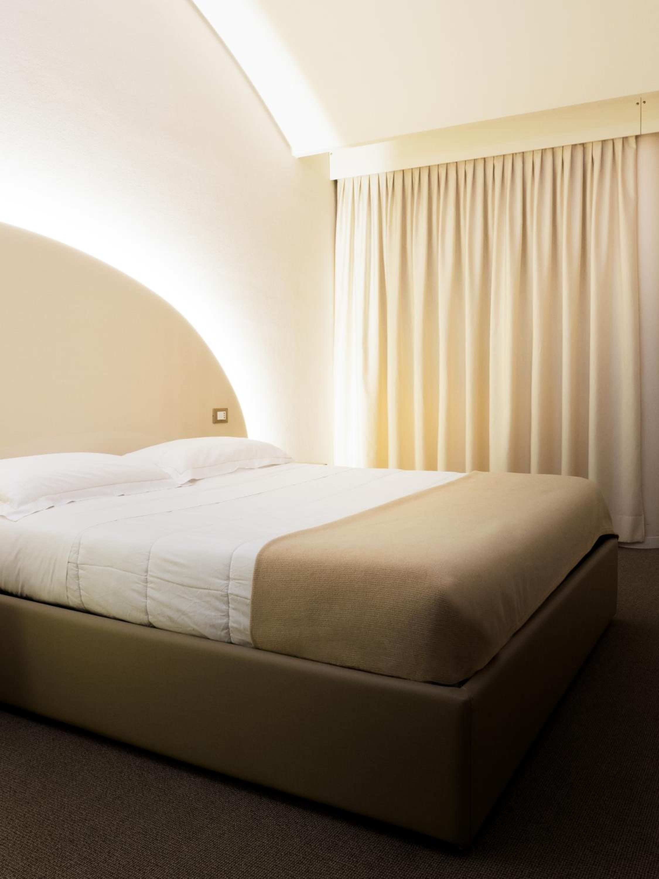 A hotel room with a single bed, a headboard, a window with curtains, and a carpeted floor.