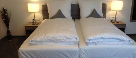 A double bed with white linens, two bedside lamps, and a vase of dried plants.