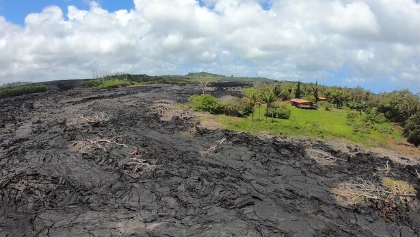 Eine Landschaft mit einem Lavafeld, einem kleinen Haus mit rotem Dach und grüner Vegetation.