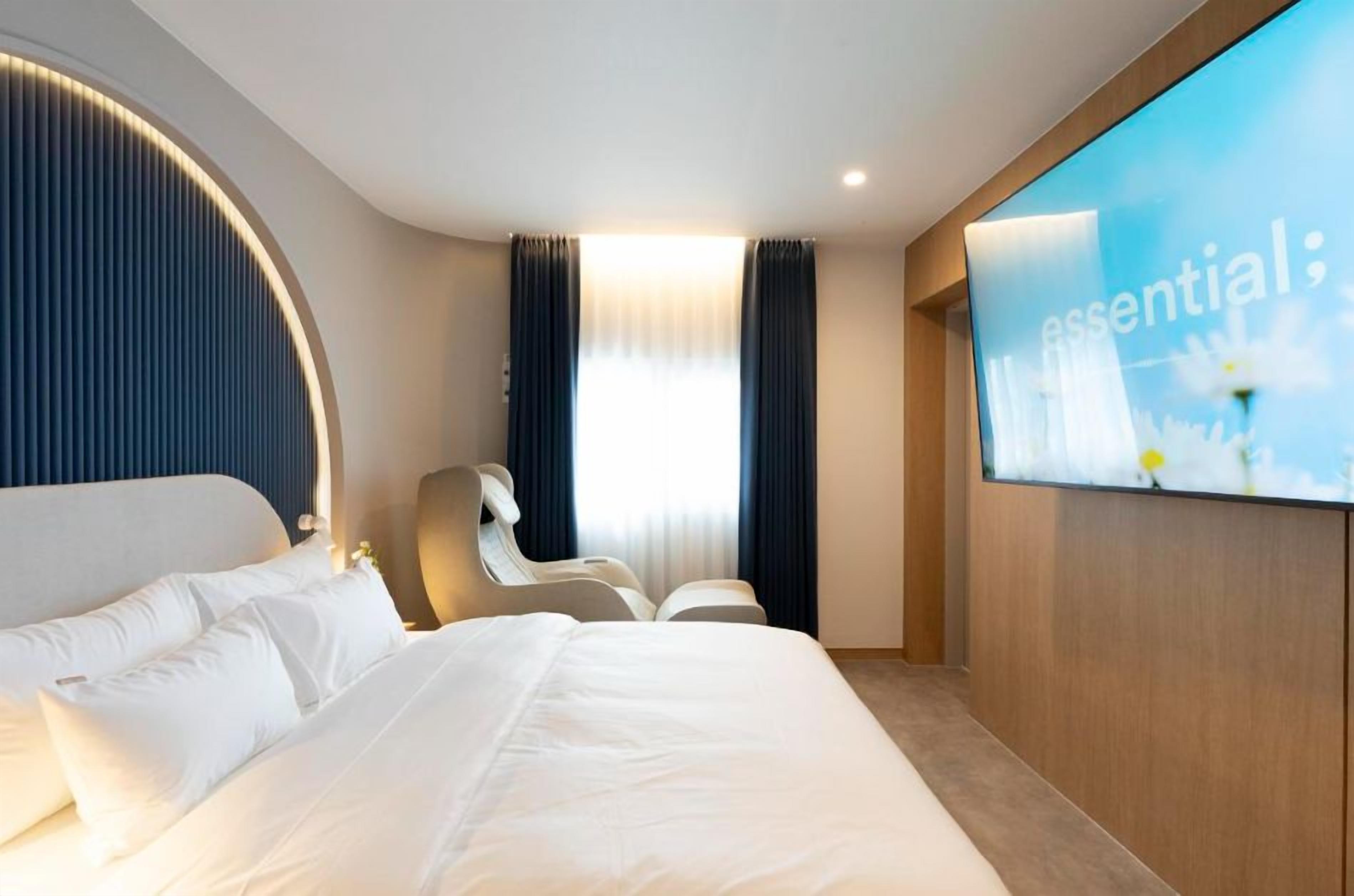 A modern hotel room with a large bed, a seating area, and a wall-mounted TV displaying the word 'essential'.