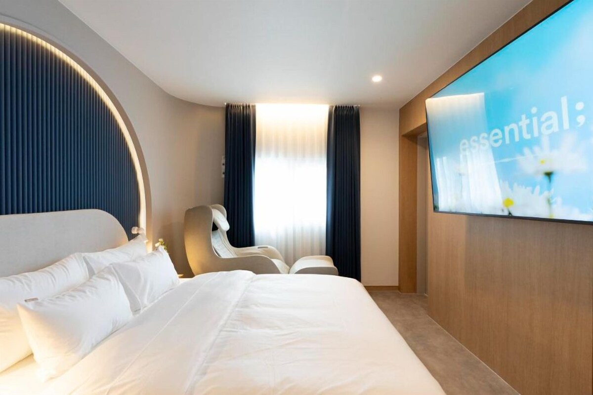 A modern hotel room with a large bed, a seating area, and a wall-mounted TV displaying the word 'essential'.