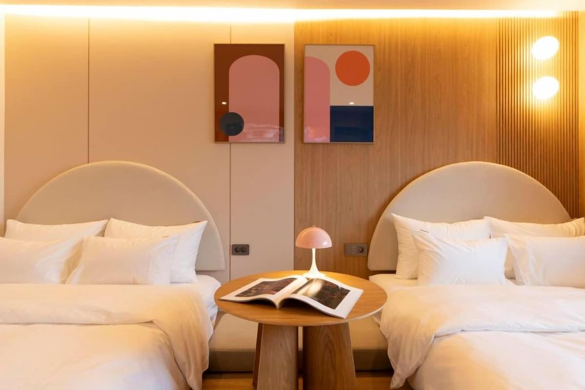 A hotel room with two beds, a small table with a lamp, and artwork on the wall.