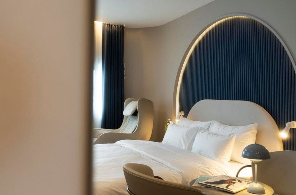 A hotel room with a curved headboard, a bed with white linens, a bedside table with a lamp, and a chair.