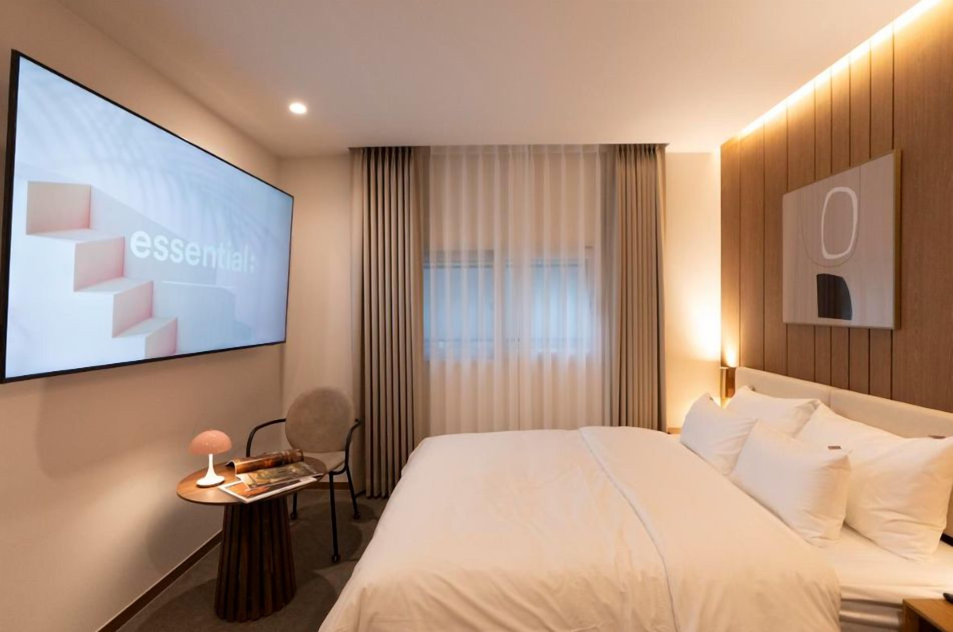 Une chambre d’hôtel avec un lit, un bureau avec une lampe, une chaise, un grand écran et une fenêtre avec des rideaux.