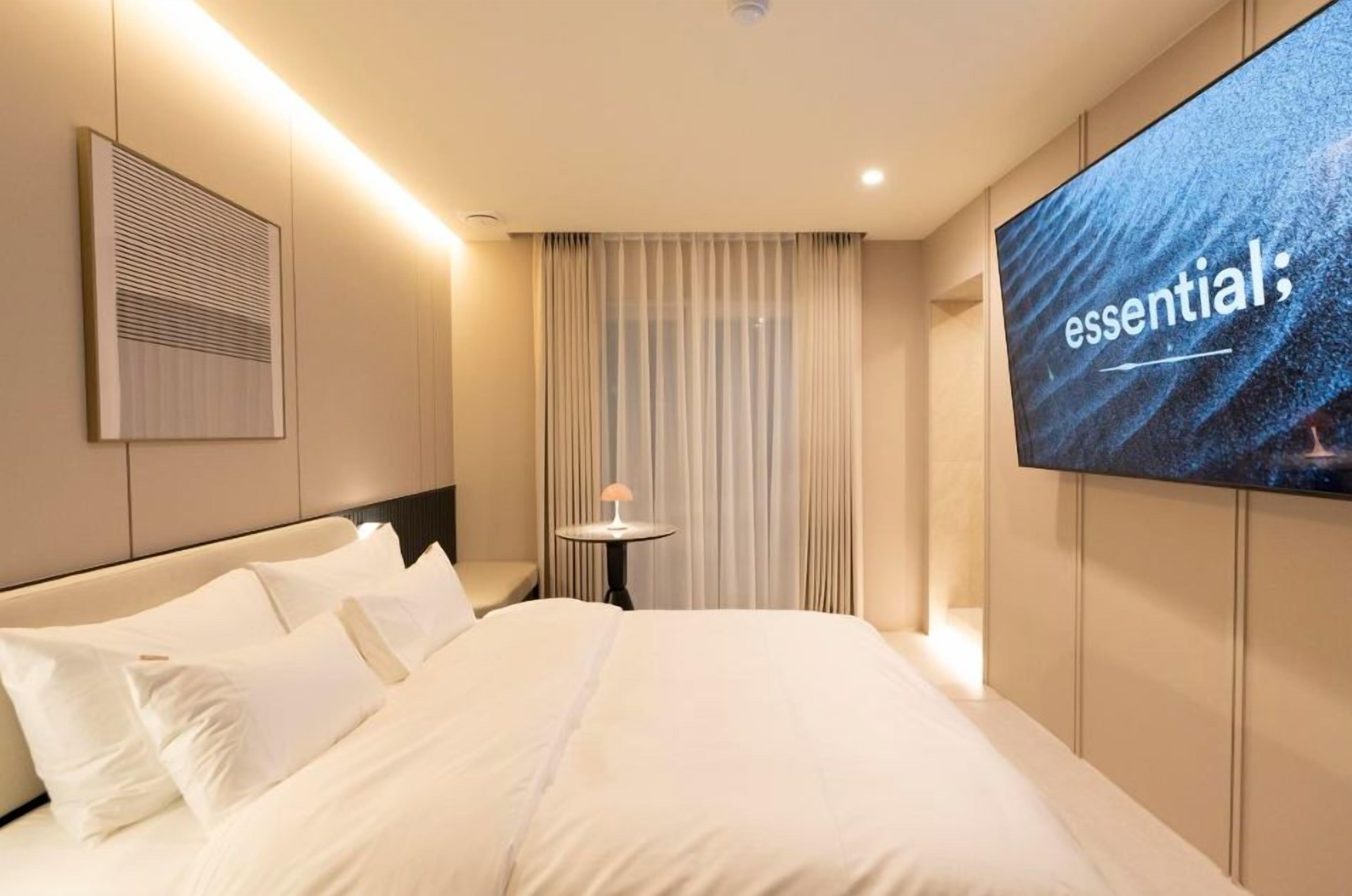 A hotel room with a large bed, a flat-screen TV mounted on the wall, a small table with a lamp, and a window with curtains.
