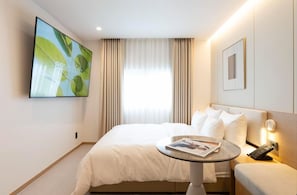 Habitación de hotel con una cama, una mesa redonda con una revista, un televisor en la pared y un ventanal con cortinas.