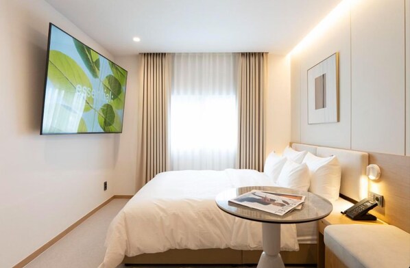 A hotel room with a bed, a round table with a magazine, a TV on the wall, and a window with curtains.