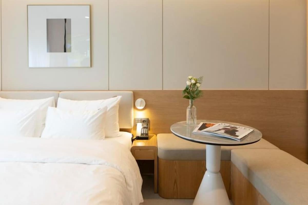 A hotel room with a bed, bedside table, and a small round table with a magazine and a vase of flowers.