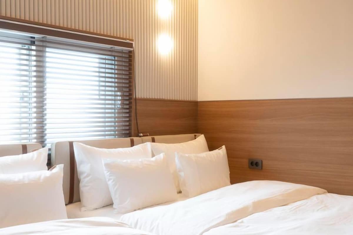 A hotel room with a bed, pillows, a wooden headboard, and a window with blinds.