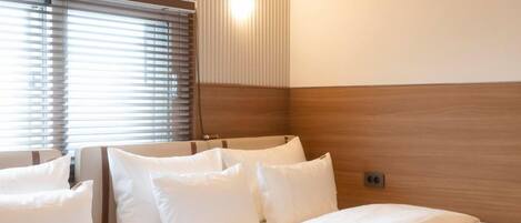 A hotel room with a bed, pillows, a wooden headboard, and a window with blinds.
