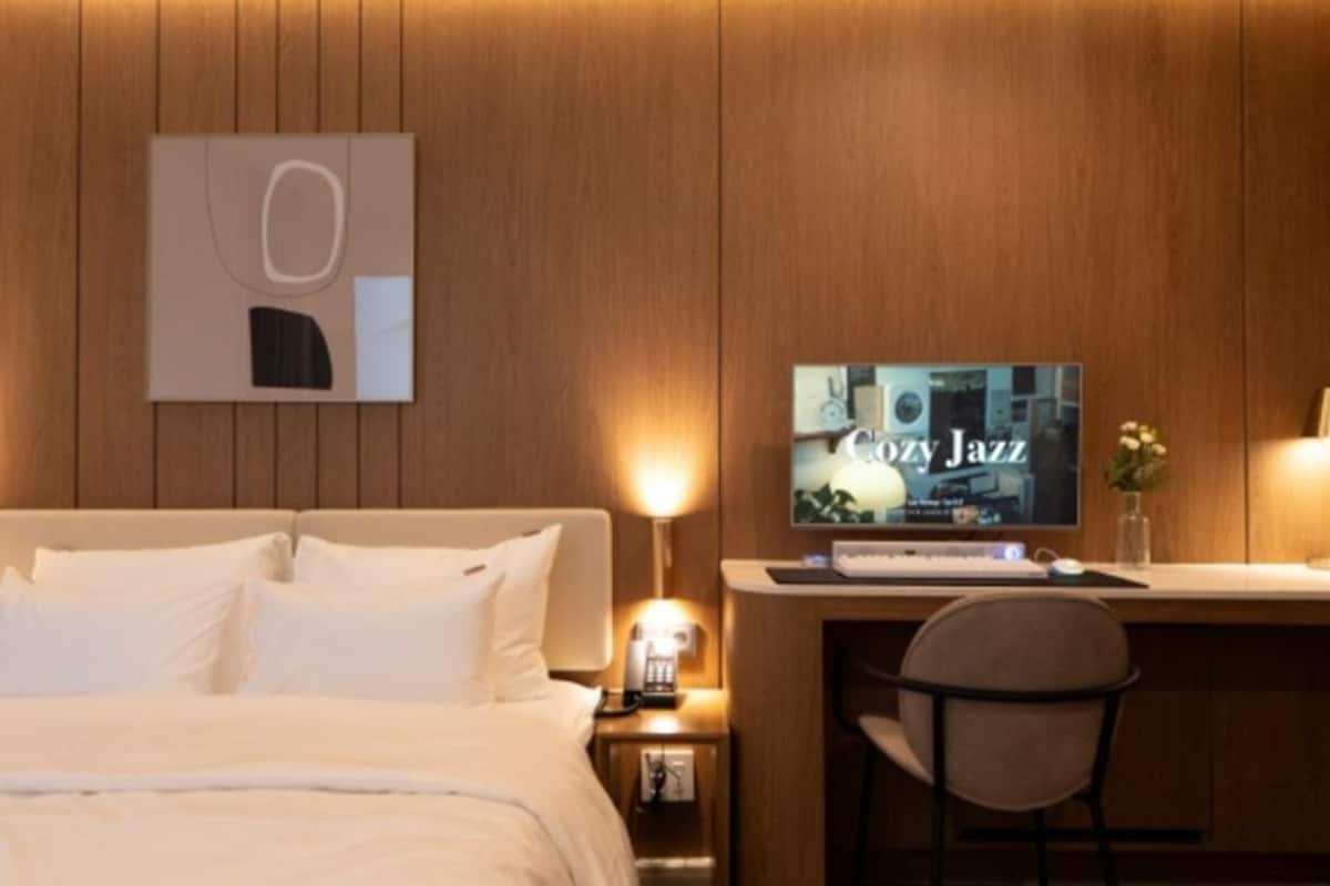A hotel room with a bed, a desk with a computer, a chair, and a television displaying 'Cozy Jazz'.